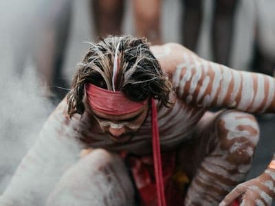 An Aboriginal man participates in a smoking ceremony, surrounded by cleansing smoke, with hands covering her face in a moment of reflection. A sacred ritual in Indigenous Australian culture, often performed to welcome visitors and protect Country.